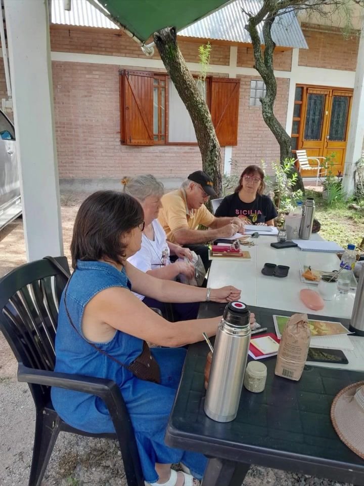 Participantes alrededor de mesas al aire libre, bajo un alero, con termos y cuadernos durante un taller.